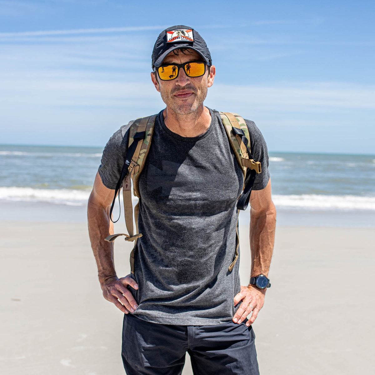 Wearing a GORUCK Spearhead Tee - Tri-Blend, a man in sunglasses, cap, and backpack stands on a sandy beach with the ocean behind him.