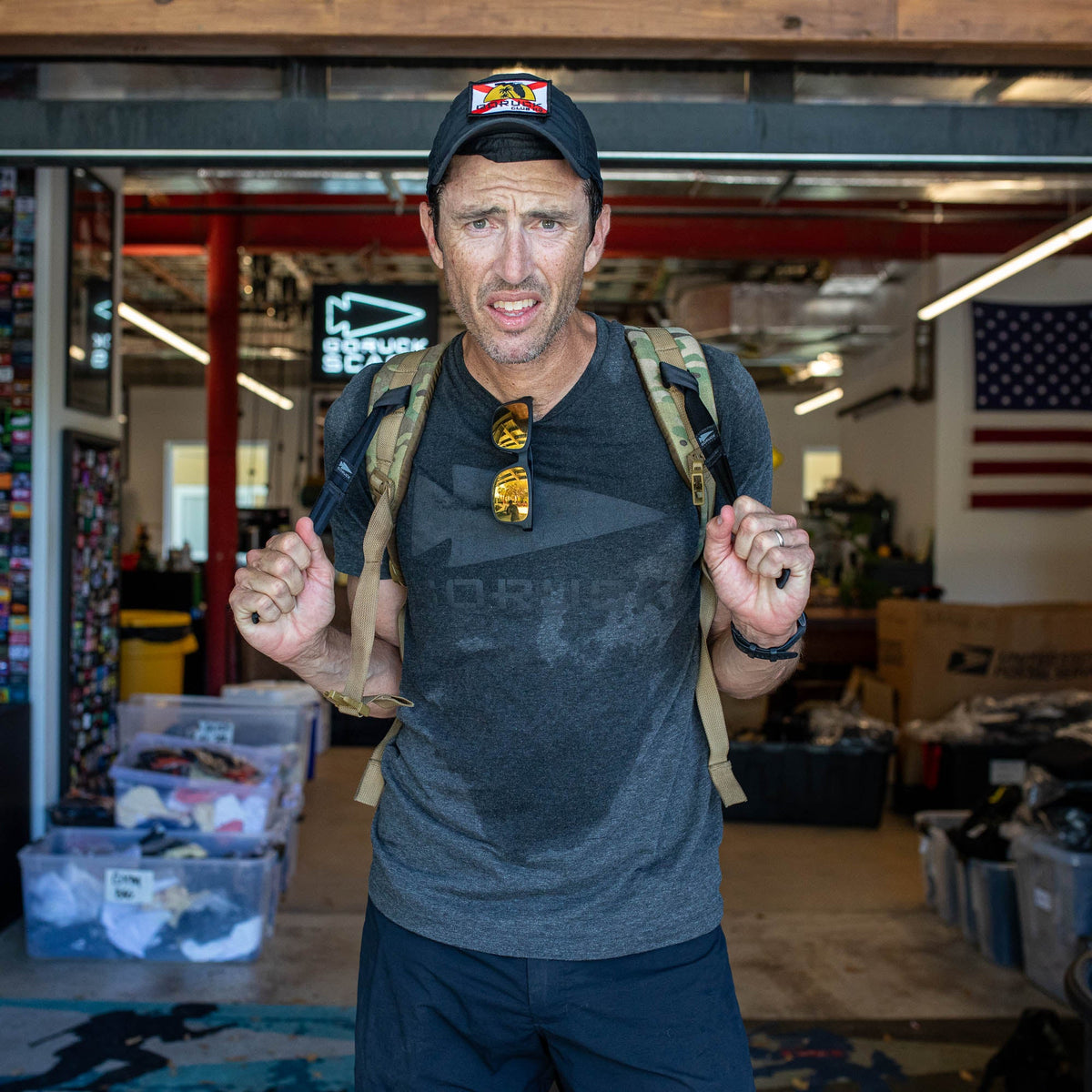 A man indoors, wearing a backpack and cap with an American flag behind him, exemplifies the grit of Special Forces and the GORUCK Spearhead spirit—now captured in the GORUCK Spearhead Tee - Tri-Blend.