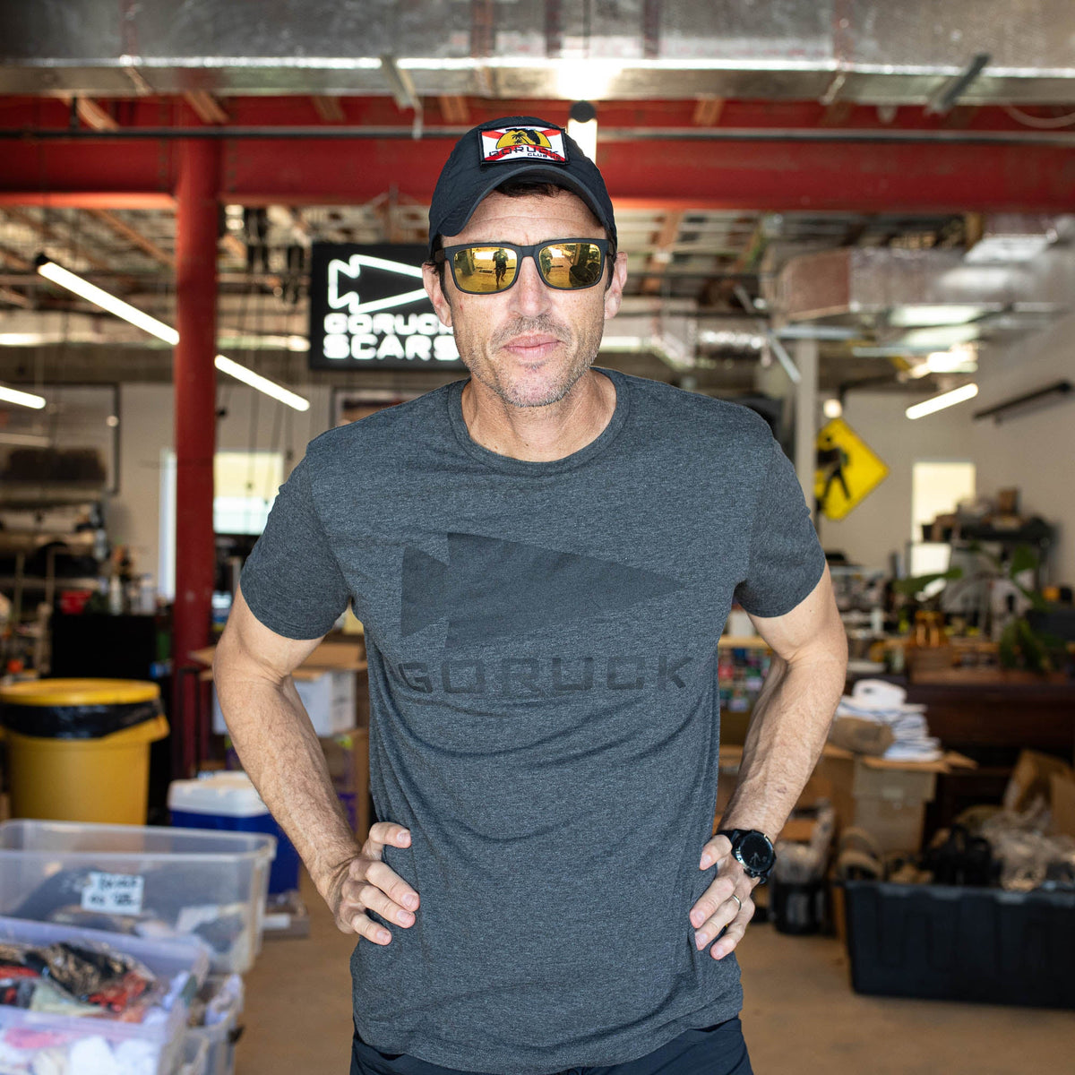 A man in sunglasses, a cap, and the GORUCK Spearhead Tee - Tri-Blend stands confidently in an industrial workshop.