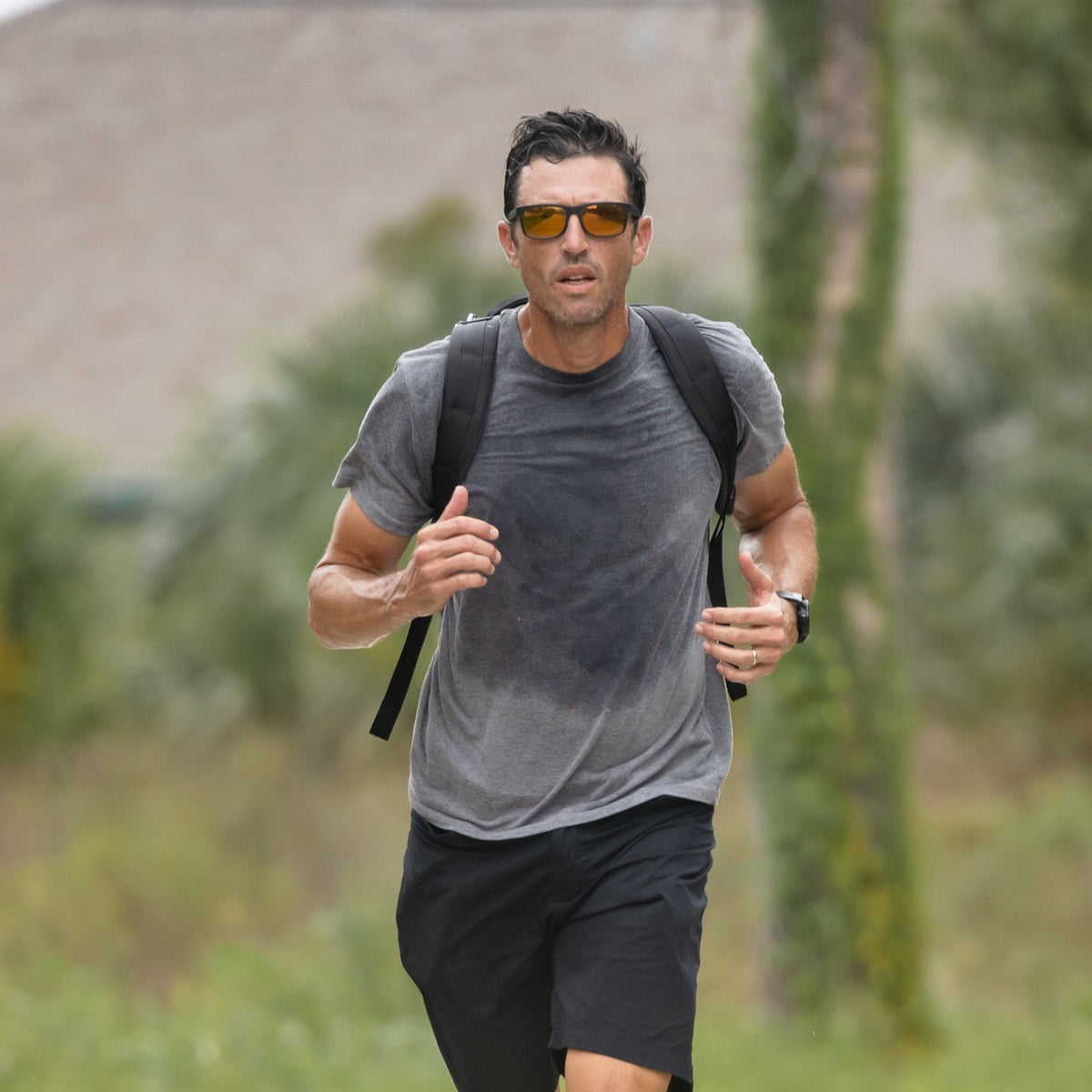 Man jogging outdoors in sunglasses and a backpack, wearing a sweaty gray t-shirt and black shorts. He carries the Basic Rucker, a rugged GORUCK rucking backpack built for endurance and adventure.