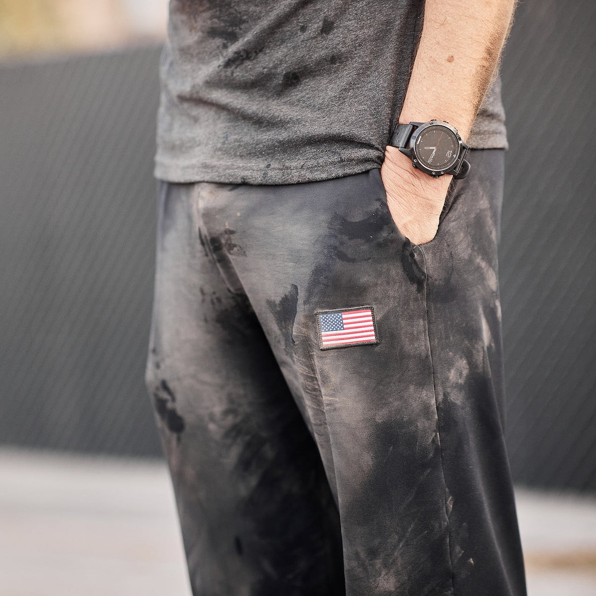 A person wearing Men’s American Training Sweatpants, featuring a tough design with an American flag patch, paired with a black watch and gray shirt visible—crafted with pride and made in the USA.