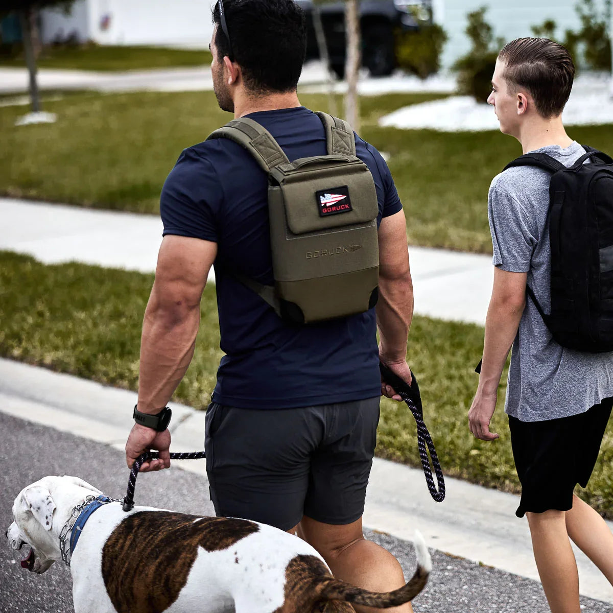 A man and teenage boy, both wearing Ruck Plate Carrier 3.0 backpacks with padded shoulder straps and ergonomic lumbar support, walk a large dog along a suburban sidewalk.