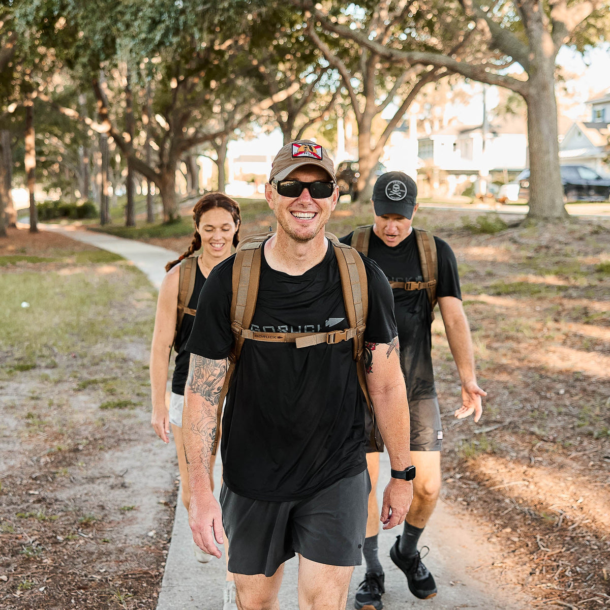 Three people walk and smile on a sunny, tree-lined path, each wearing backpacks equipped with the Training Sternum Strap for adjustable weight distribution.