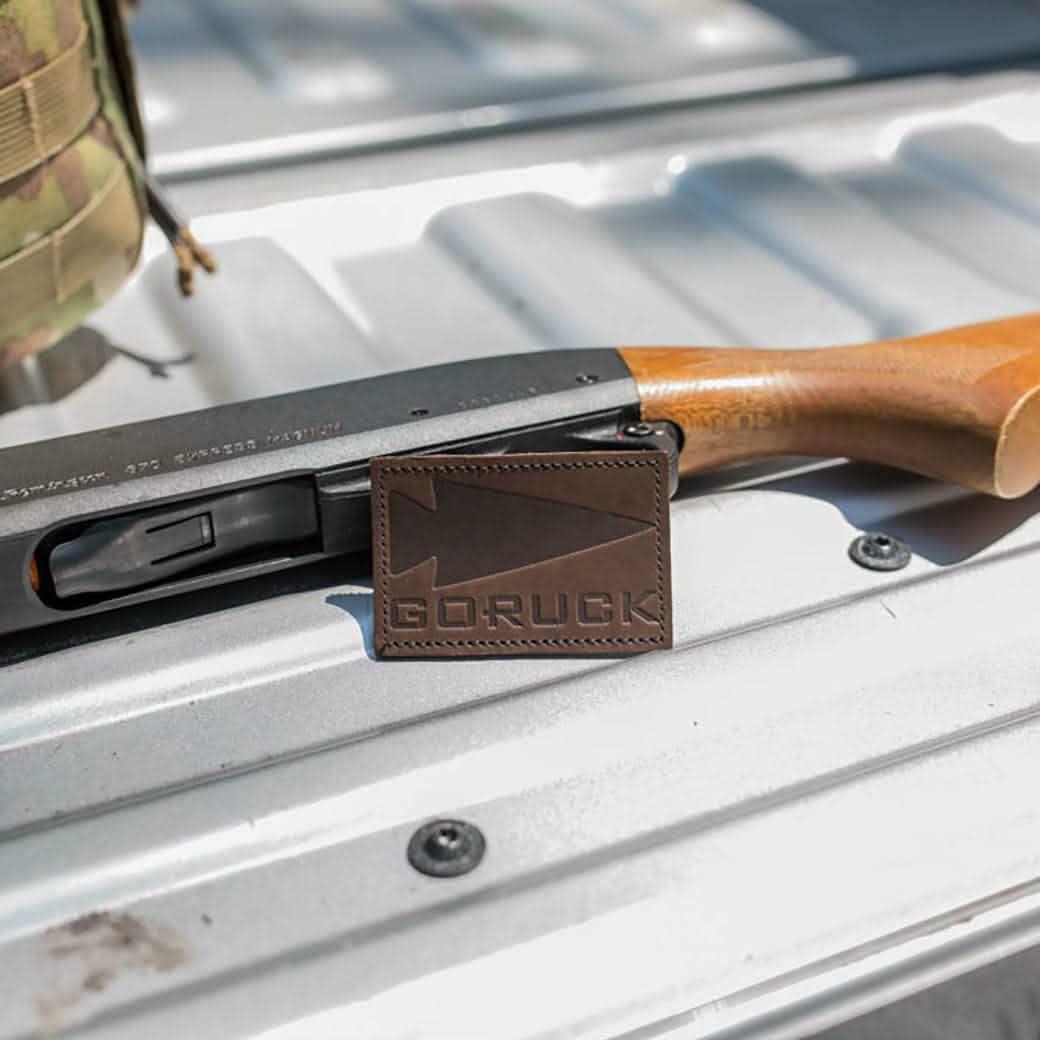 GORUCK leather patch with arrowhead logo on metal surface, shotgun and backpack nearby