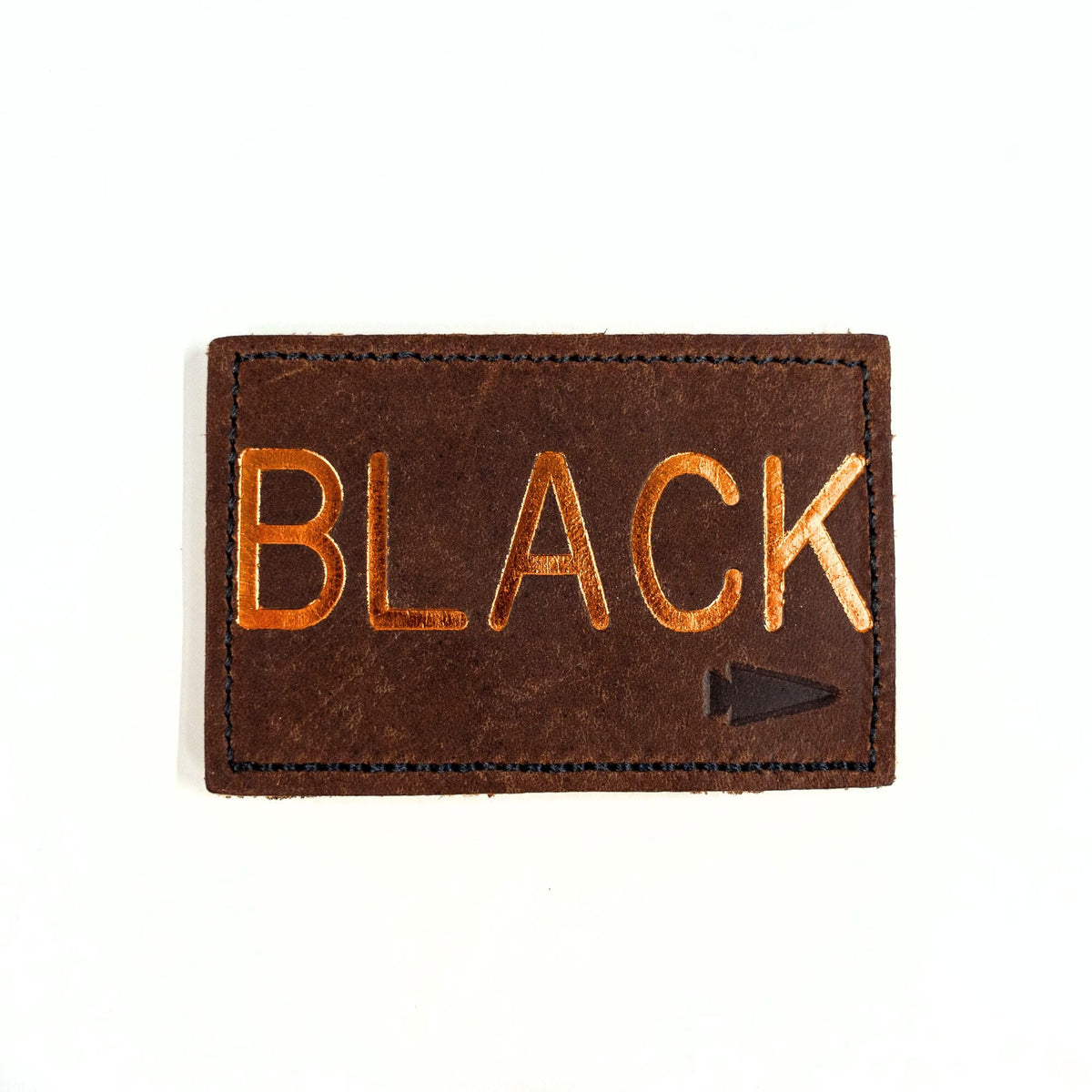The Patch - Custom Leather Monogram is a brown leather patch, personalized with "BLACK" embossed in gold and accented with a small arrow symbol in the corner.