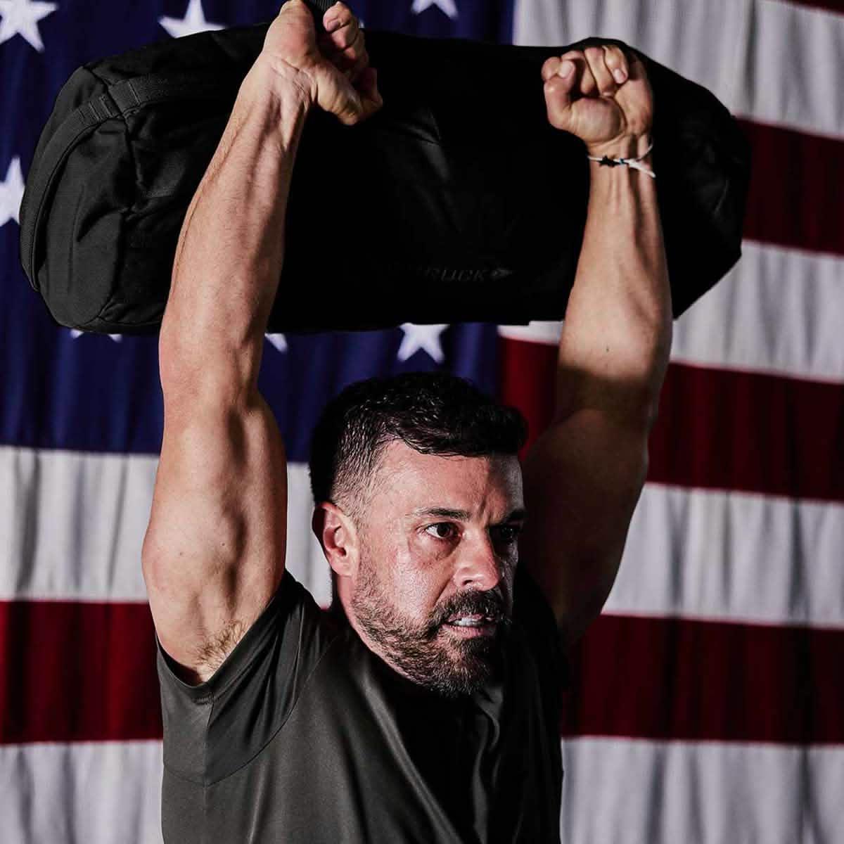 Man lifting GORUCK sandbag overhead, American flag in background, rucking workout gear.