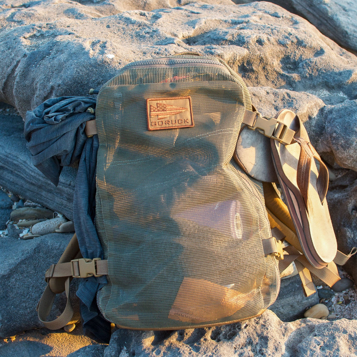 The Mesh Ruck backpack with a GORUCK patch, sandals, and clothing sits on sunlit rocks, drying quickly thanks to its breathable mesh design.
