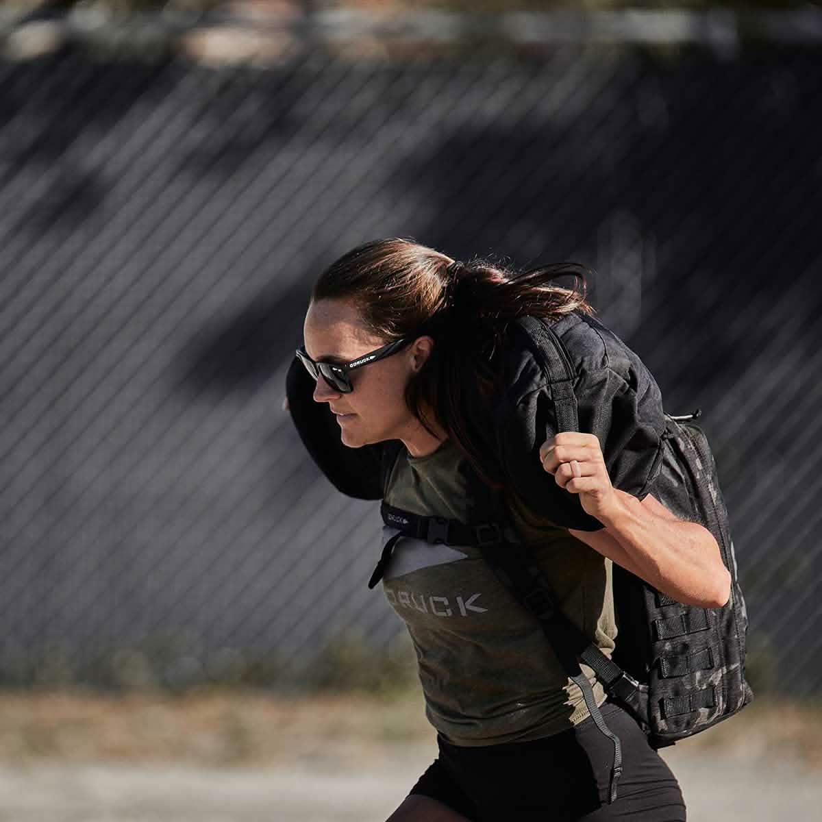 Woman carrying GORUCK 80lb sandbag outdoors, wearing sunglasses and GORUCK shirt
