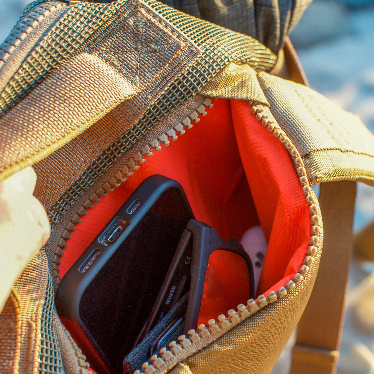 A close-up of an open tan Mesh Ruck pocket shows a smartphone, a case, and white earbuds inside.