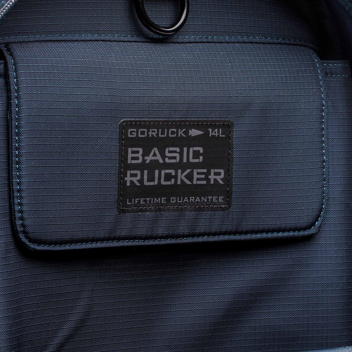 Close-up of the front pocket on a blue Basic Rucker backpack, featuring a stitched label and designed for comfortable rucking with secure storage for your ruck plate.