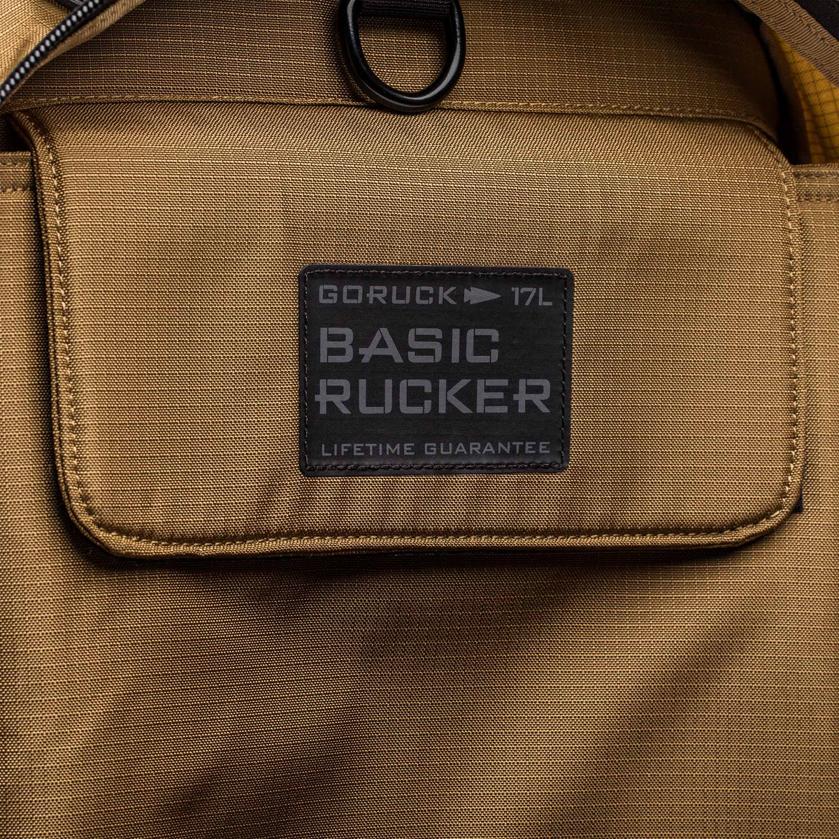 Close-up of a tan Basic Rucker® backpack pocket, featuring a label that reads "17L BASIC RUCKER LIFETIME GUARANTEE.