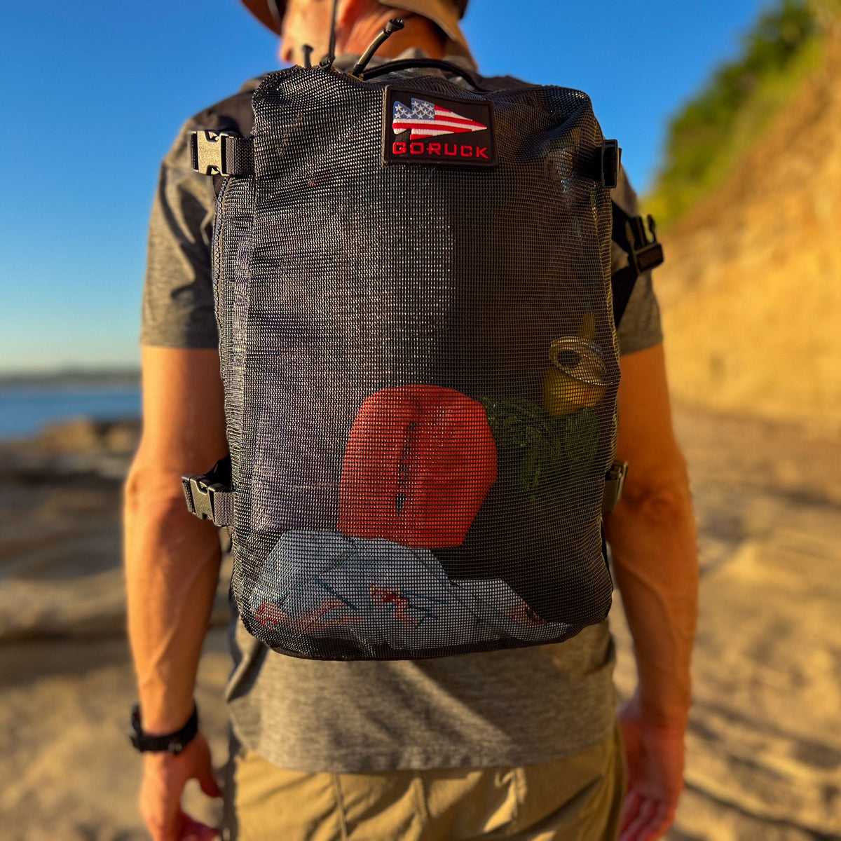 A person carries the Mesh Ruck, a black mesh backpack with visible gear inside, while walking outdoors on a sunny day.