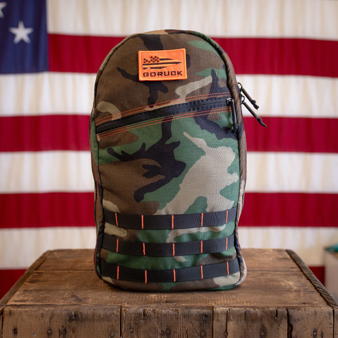Woodland Camo 15L Bullet ruck with an orange morale patch, standing on a wooden crate in front of an American flag.