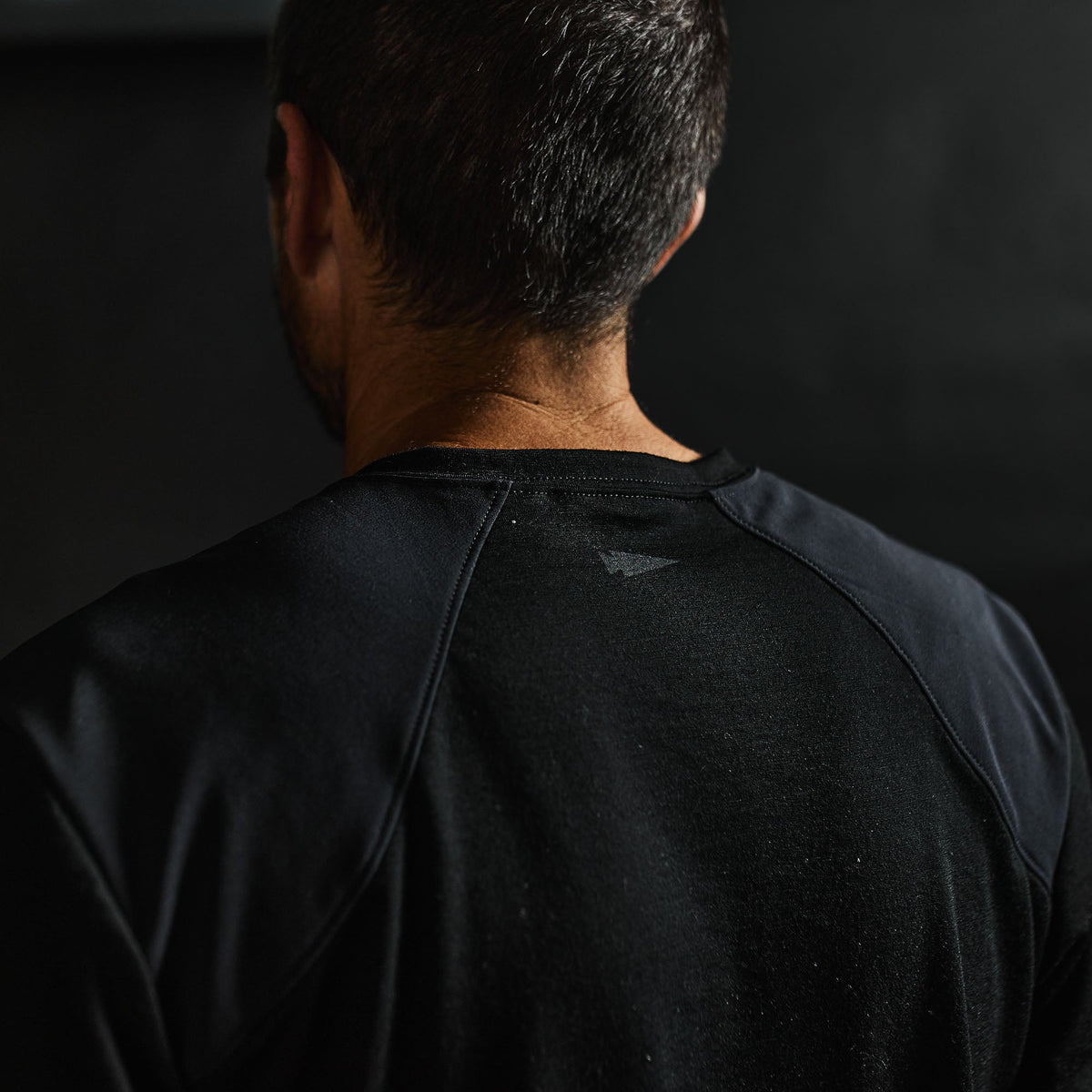 Close-up of a man’s back wearing the Men’s Commando Tee - Merino Wool in black, featuring reinforced shoulders. His short dark hair stands out against the dark background.