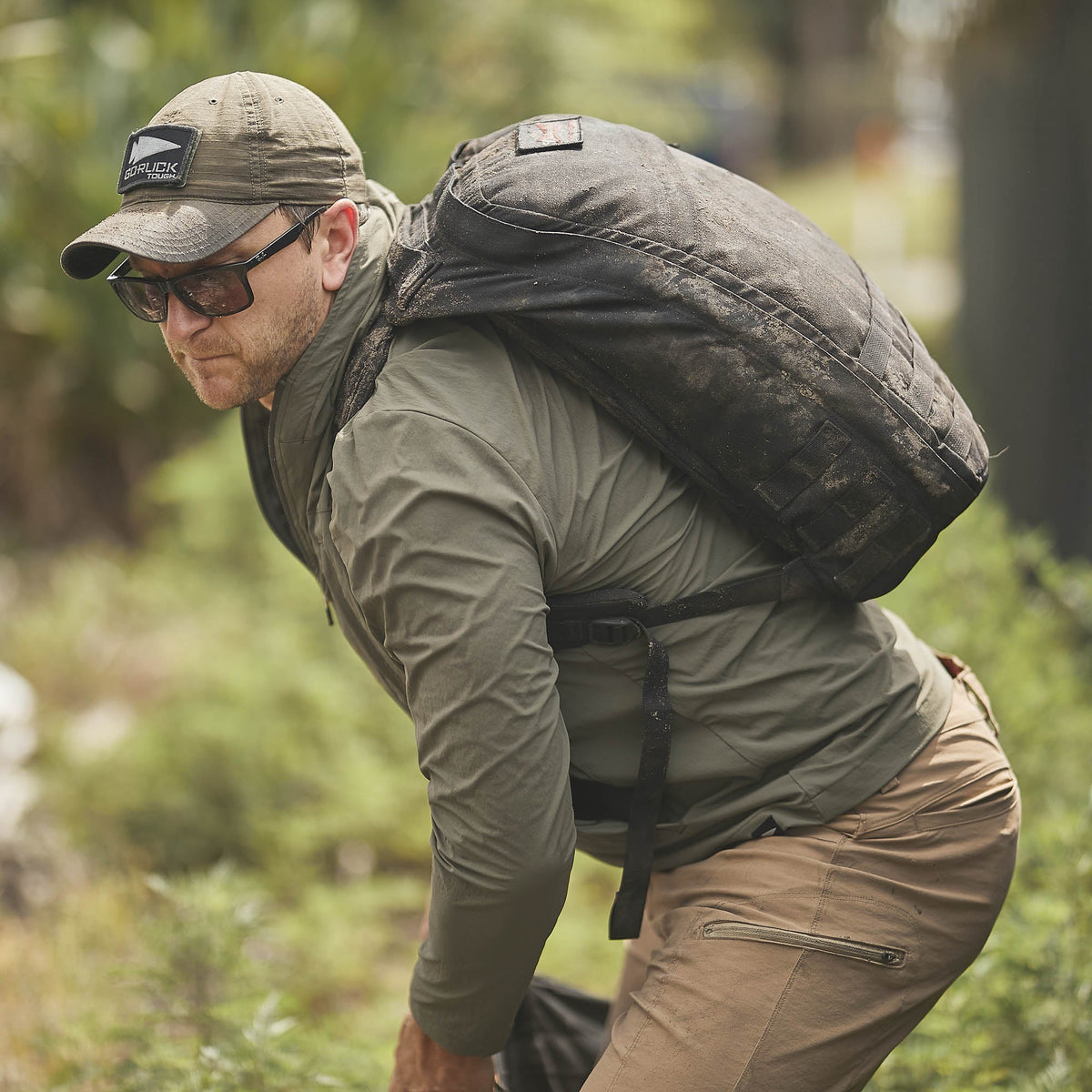 Man in outdoor gear and sunglasses, carrying a large backpack in a green setting, wears the abrasion-resistant Indestructible Challenge Windbreaker designed for rugged adventures.