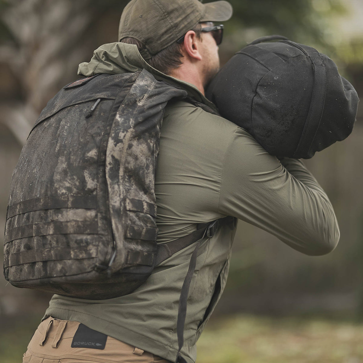 Man in outdoor gear carrying a sandbag and wearing a dirty black backpack, dressed in an Indestructible Challenge Windbreaker made from abrasion-resistant ToughDry fabric.