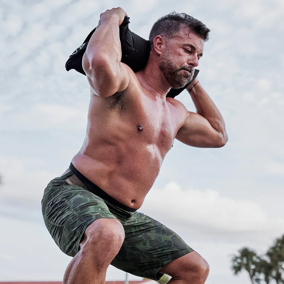 Shirtless man doing outdoor squat with weighted ruck, wearing camo shorts, GORUCK fitness gear