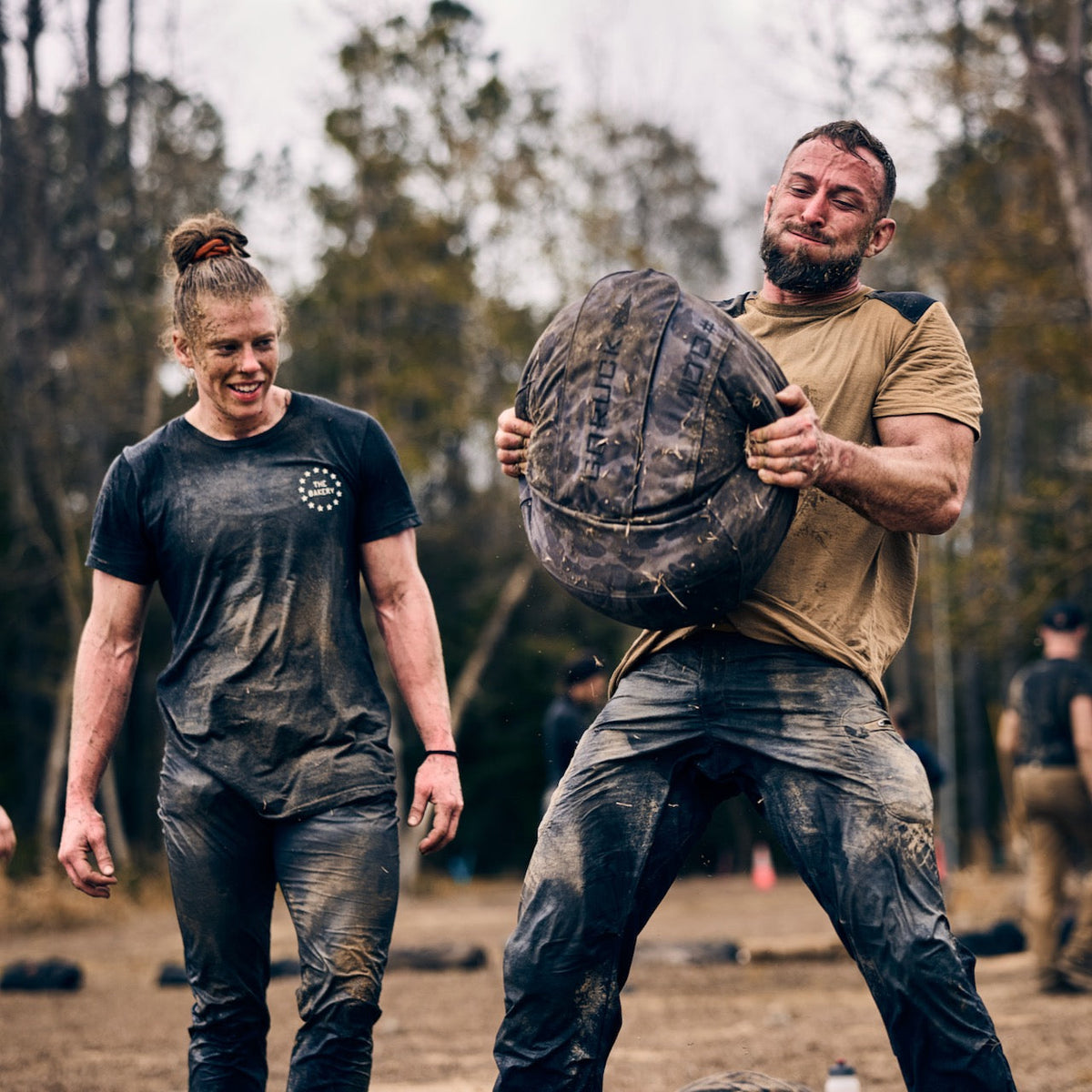 A man in abrasion resistant pants strains to lift a heavy sandbag as a woman watches him during a muddy obstacle course race.