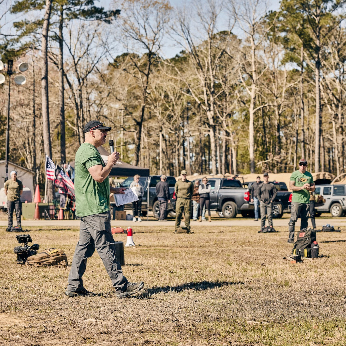 A man in a green t-shirt and ToughDry pants speaks into a microphone while holding papers, addressing a group of people standing in a field in front of parked trucks and trees.