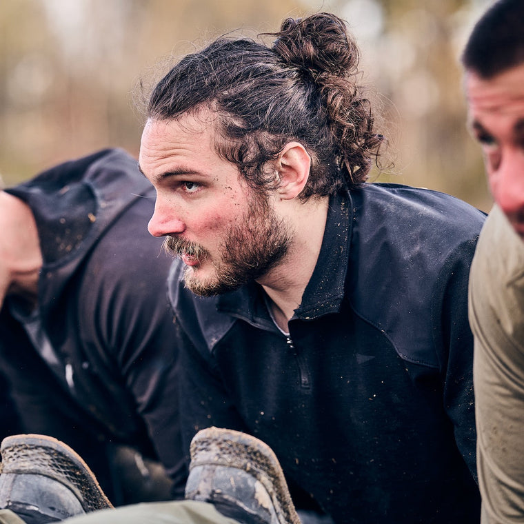 A man with curly hair in a bun and a black Men's Commando Quarter Zip - Merino Wool jacket leans forward outdoors, looking focused.