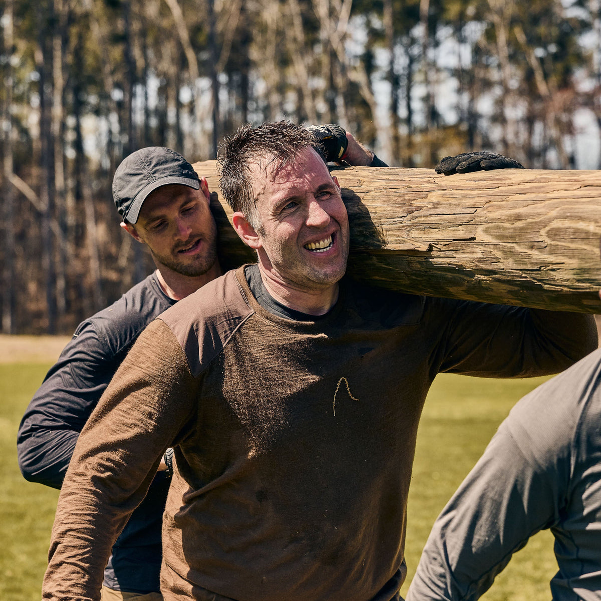 Two men outdoors, both wearing the Men’s Commando Long Sleeve - Merino Wool shirts with reinforced shoulders, team up to carry a large wooden log during a physical challenge.