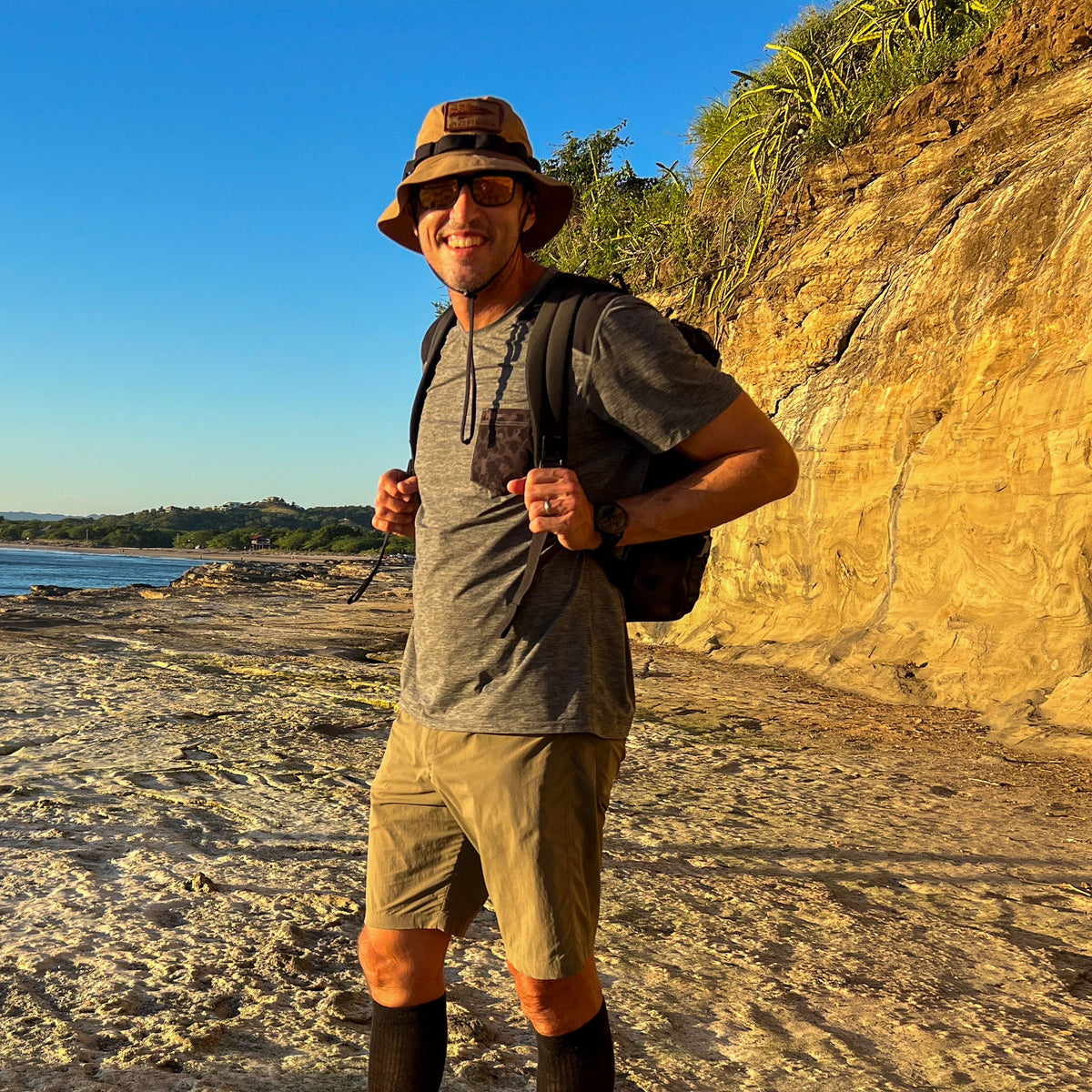 A man in a hat and sunglasses smiles on a rocky shore with cliffs and blue sky, wearing the Mesh Ruck, a quick-drying backpack.