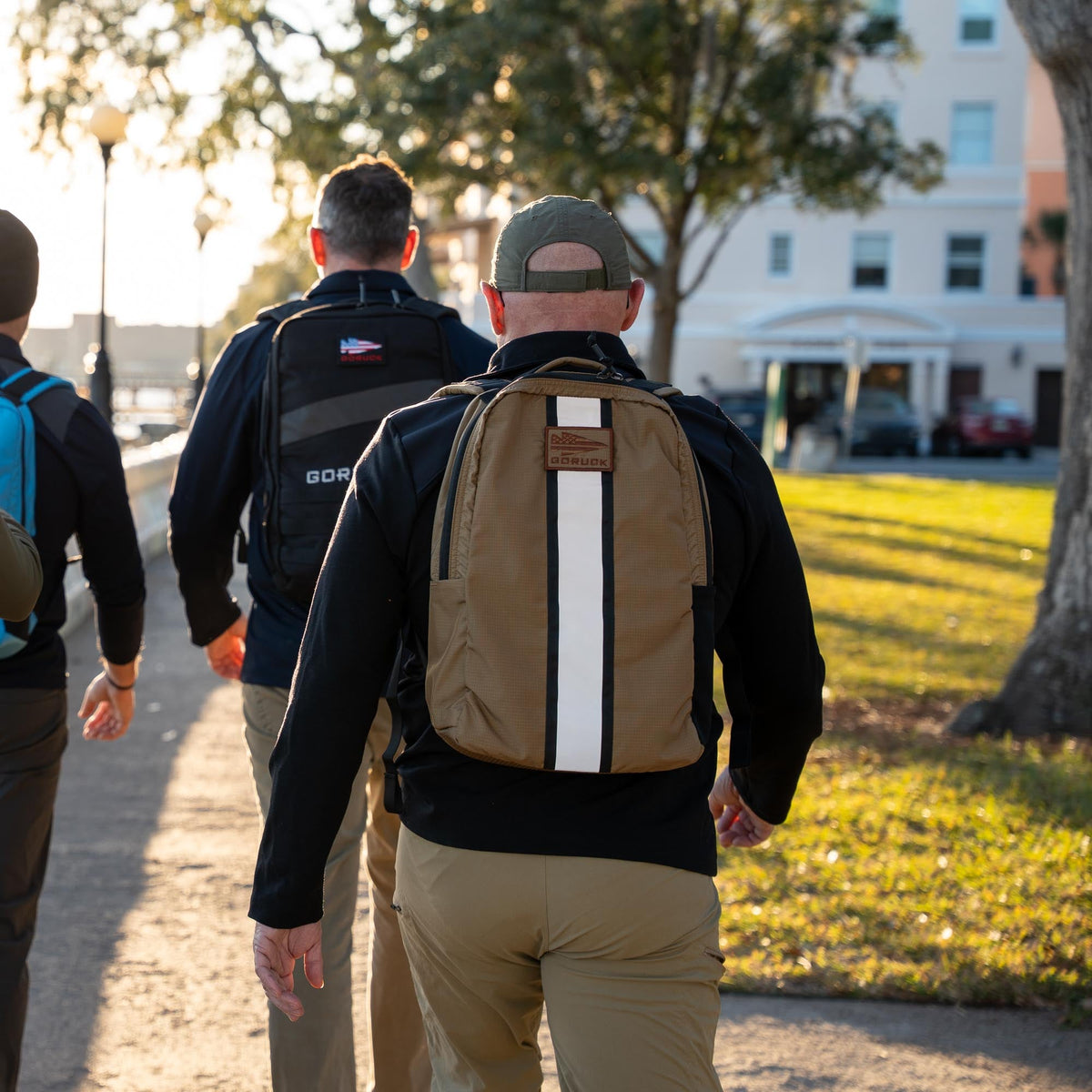 Three people with backpacks, including the Basic Rucker®, walk on a sunlit park path with trees and a building ahead.