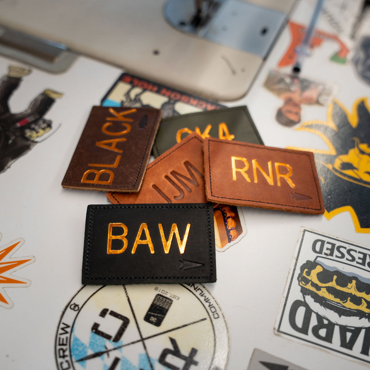 Patch – Custom Leather Monogram features personalized leather patches with gold lettering, displayed on a table with colorful stickers and labels—ideal for gifts or adding custom initials to your favorite items.