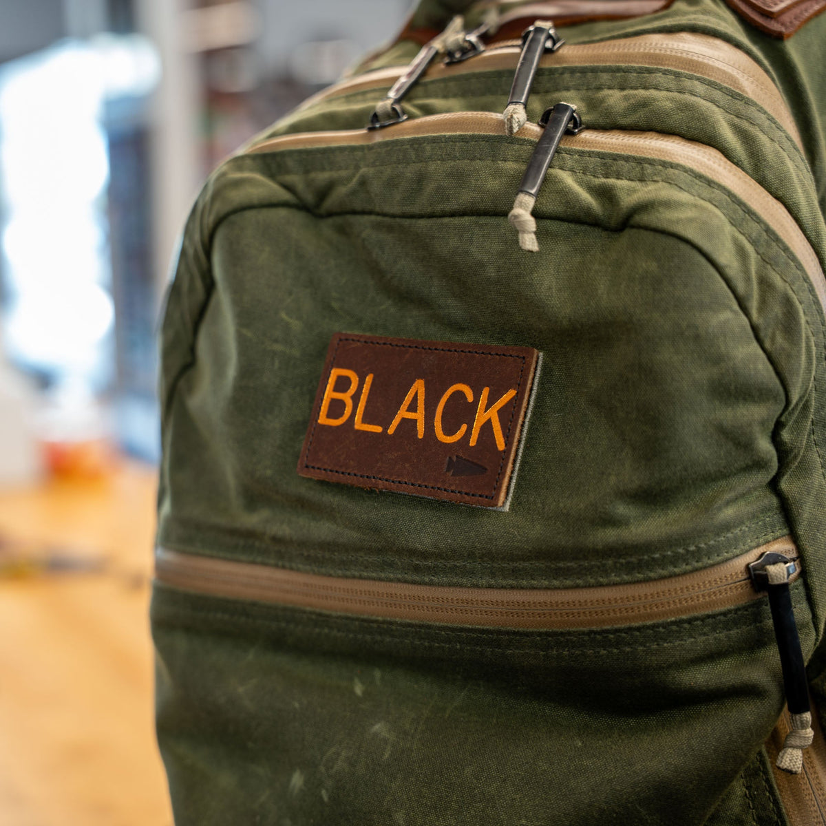 A green backpack features a brown Custom Leather Monogram Patch labeled "BLACK" on the front pocket.