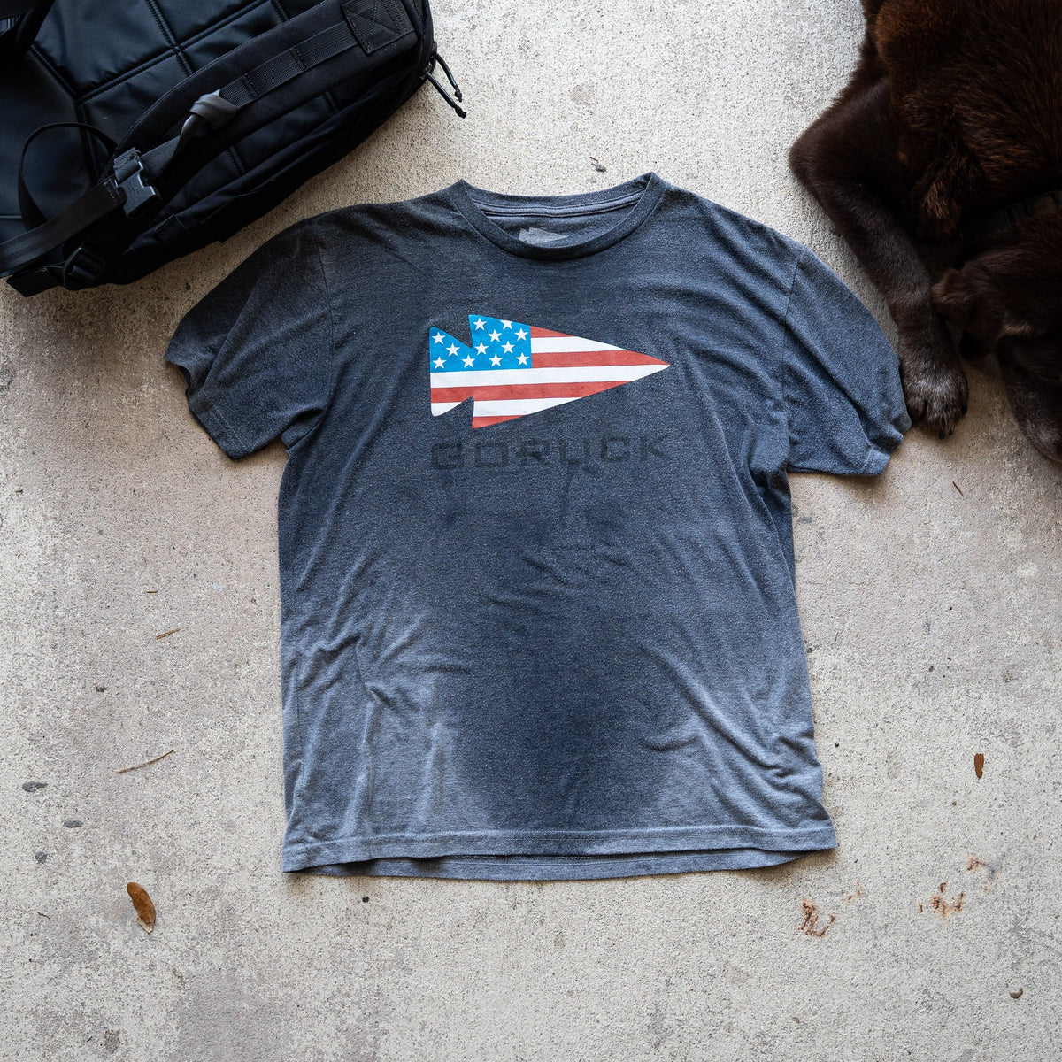 The GORUCK Flag Spearhead Tee - Unisex Tri-BlendX, a gray premium T-shirt featuring an American flag and Special Forces-inspired spearhead, is shown on concrete next to a black bag and a dog's paw.
