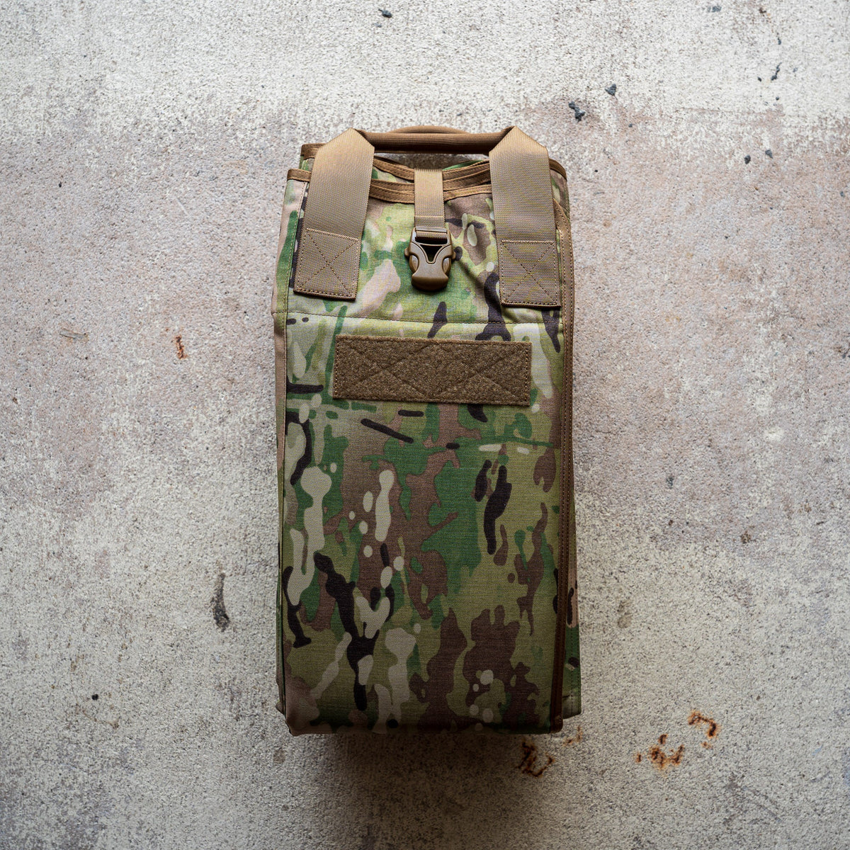A folded camouflage Shooting Mat with tan straps rests on a textured concrete surface.