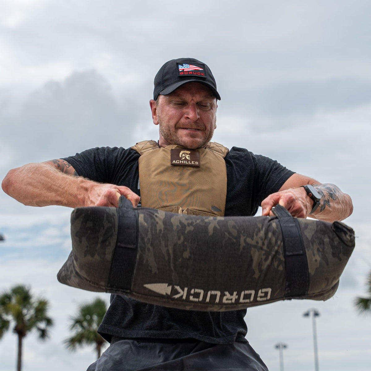 A man wearing the Achilles | Men's Weighted Vest lifts a heavy sandbag outdoors with palm trees and a cloudy sky in the background.