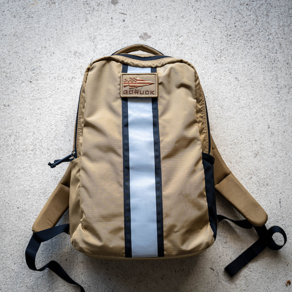 The Basic Rucker backpack, featuring a tan design with a reflective gray stripe, a GORUCK patch, and space for a ruck plate, rests on a light concrete surface—ideal for your next rucking adventure.