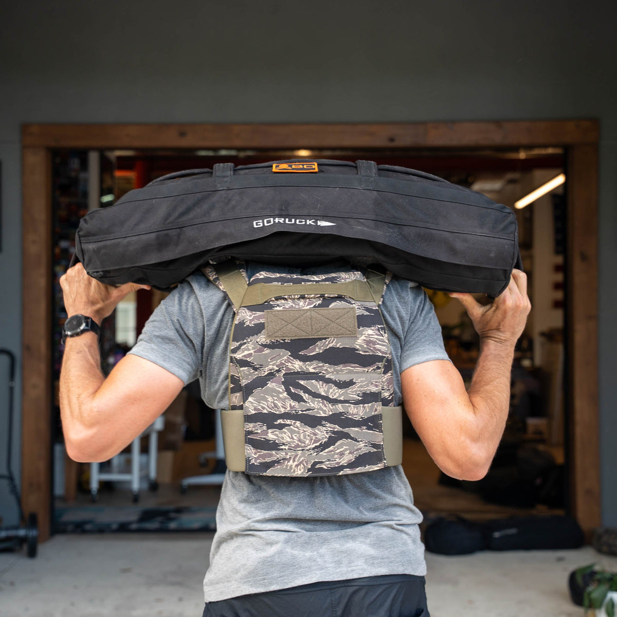 Person lifting a black GORUCK sandbag onto their shoulders, wearing the Rucking Weight Vest in a camo pattern.