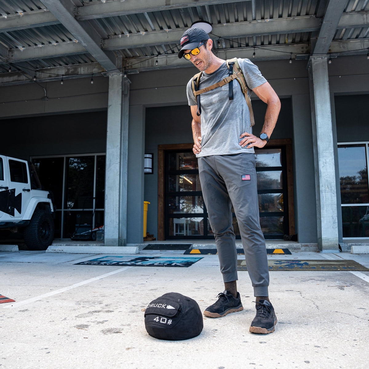A man in athletic gear stands outside, looking down at a weighted sandbag on the ground in front of him, confidently dressed in Men’s American Training Sweatpants—made in the USA and built to last with a lifetime guarantee.