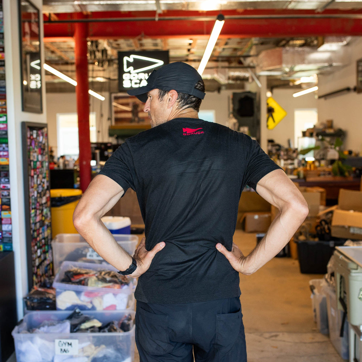 A man wearing the GORUCK Spearhead Tee - Tri-Blend in black and a cap stands hands on hips, facing away in a cluttered indoor workspace.