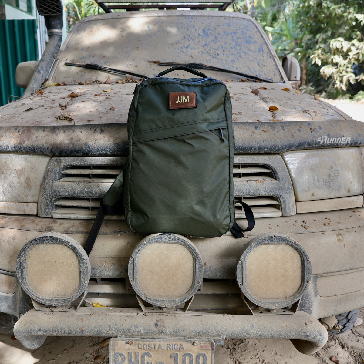 A green backpack featuring a Patch - Custom Leather Monogram sits on the dusty hood of a dirt-covered truck with Costa Rica plates.