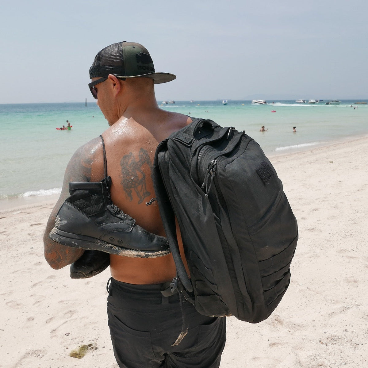 A man on a sandy beach carries the GR2 USA - Cordura travel backpack and boots, with people swimming and boats visible in the background.