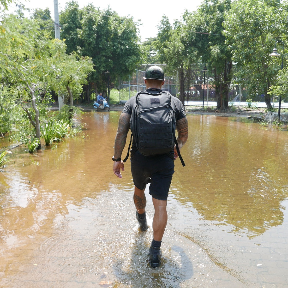 A man carrying a GR2 - Cordura ruck walks along a flooded path lined with trees on a sunny day.