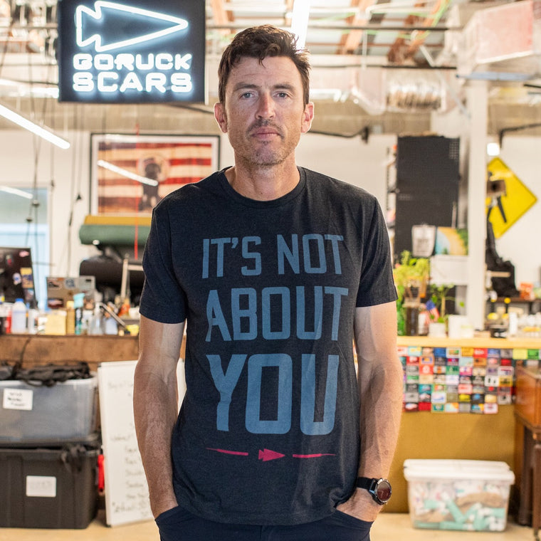 A man wearing the “It’s Not About You - Tri-BlendX - Unisex” shirt stands in an office with industrial decor and signs. This premium Tri-BlendX shirt, printed in the USA, combines style and comfort for everyday wear.