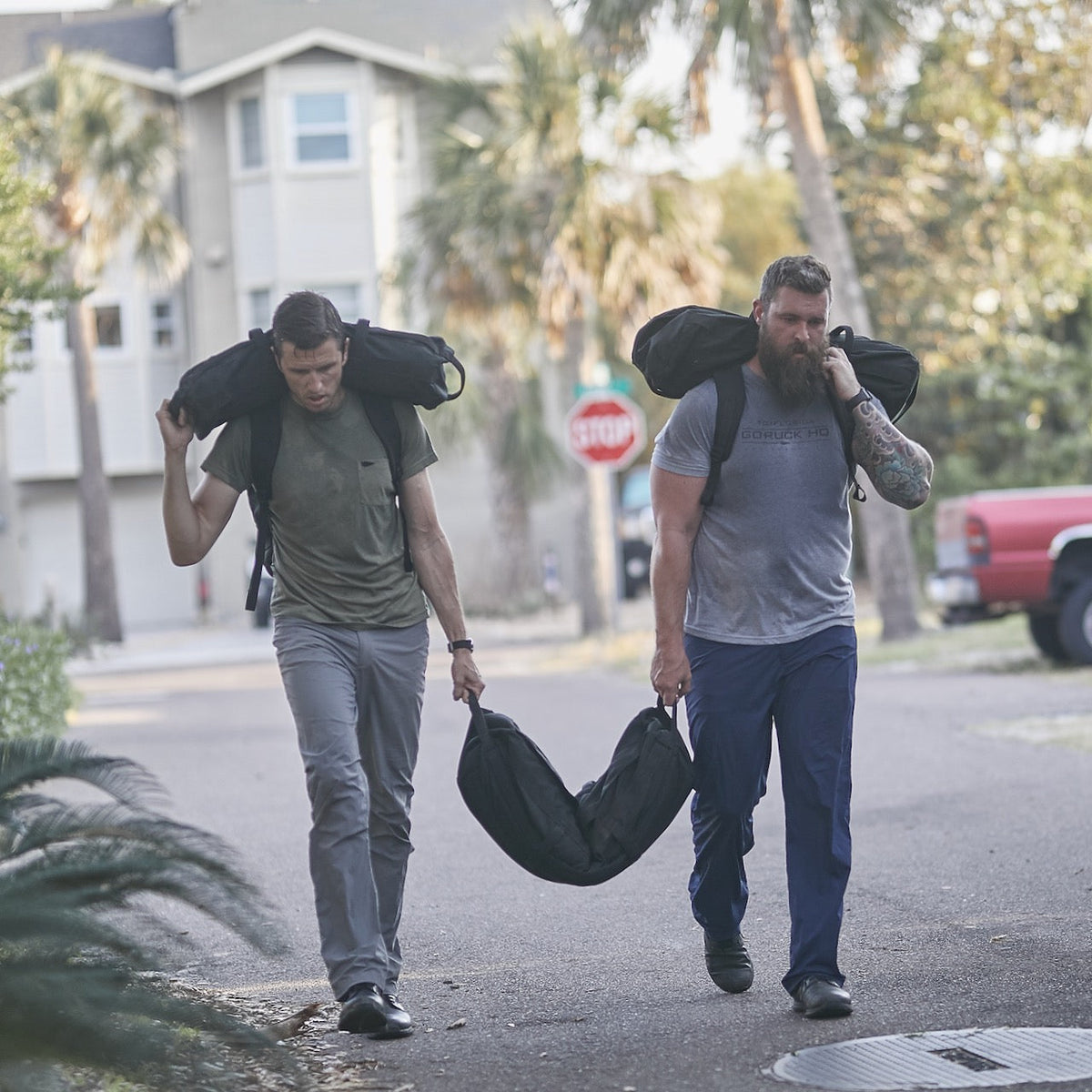 Two men carry Sandbag Kit (w/ Filler Bag) on their shoulders while walking down a residential street, pushing their limits with these durable training sandbags built to last.
