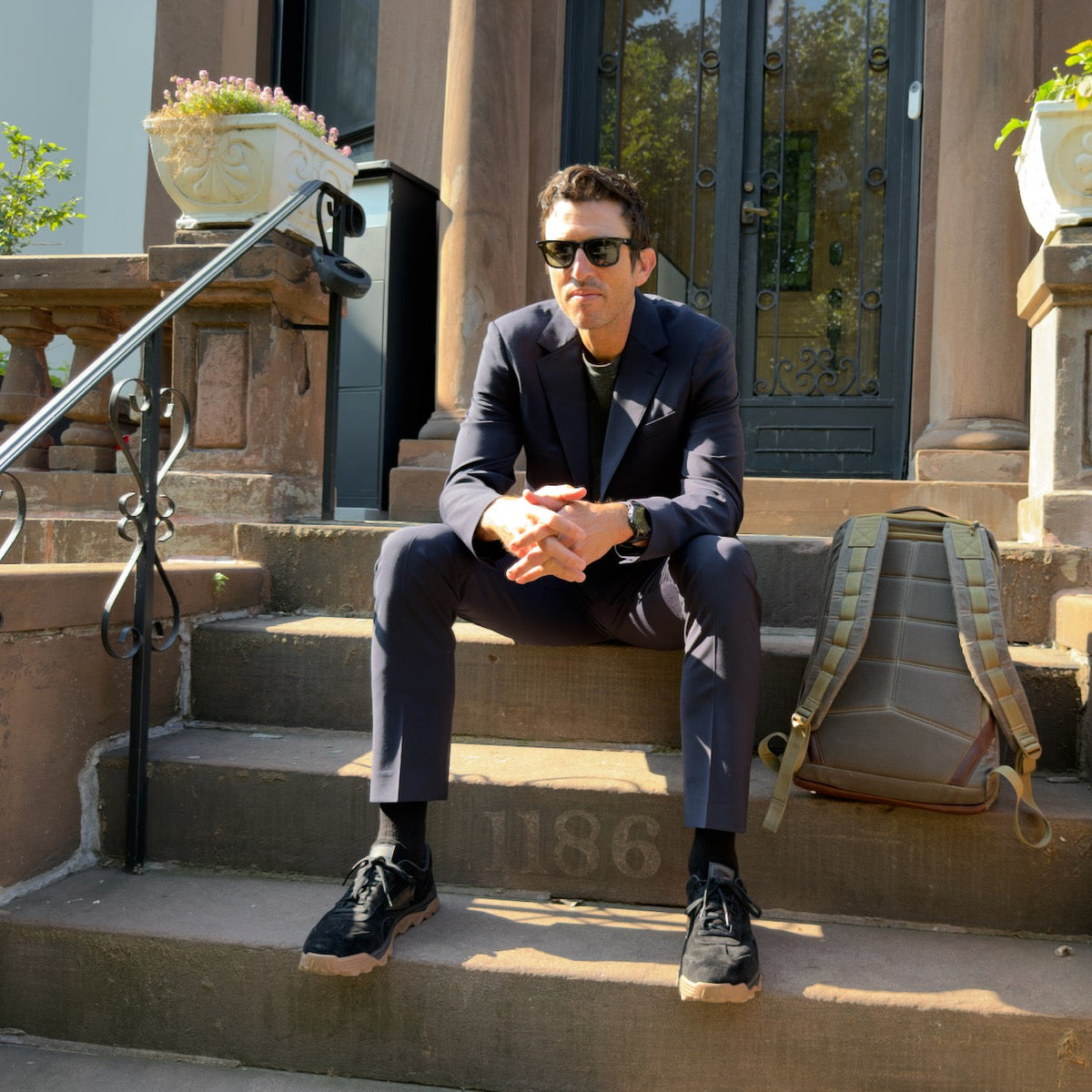 A man in sunglasses and a suit sits on stone steps by a backpack, wearing Mackall 45 Black + Gum sneakers, in front of a brownstone building.