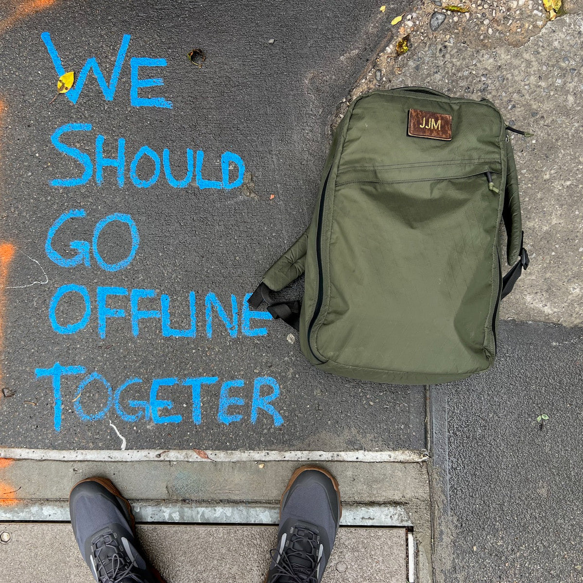 Patch - Custom Leather Monogram backpack in olive green rests on pavement with a personalized leather patch. Blue chalk text says, "We should go offline together." Feet are visible at the bottom.