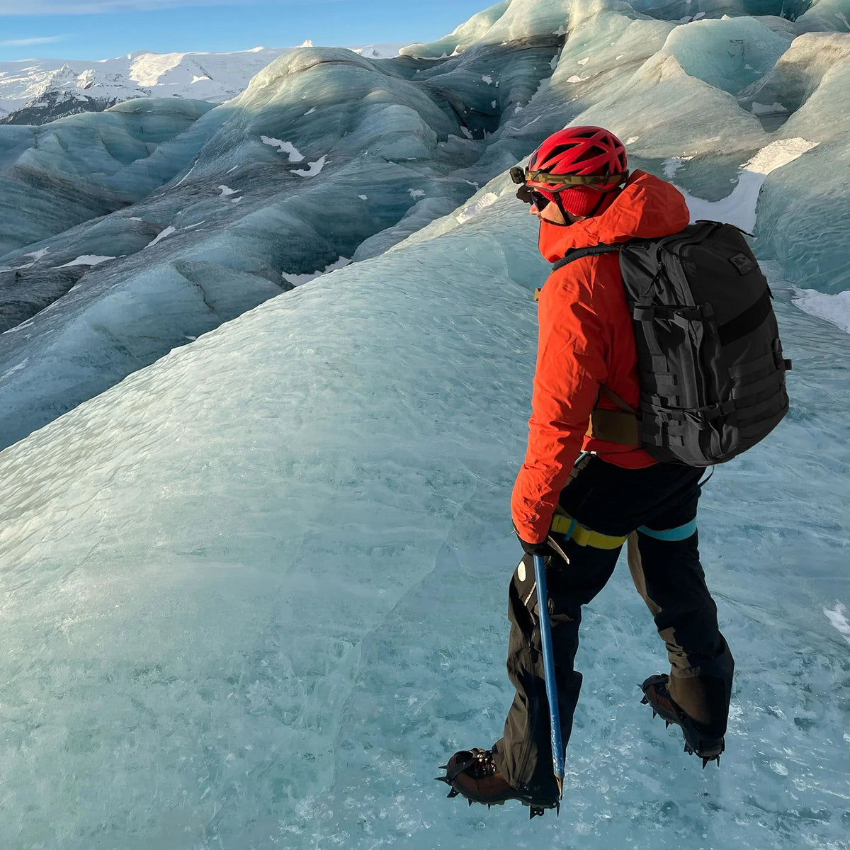 Wearing a red jacket and helmet, a person stands on a glacier with climbing gear and a Rucker - Long Range rucksack, gazing over the icy landscape.