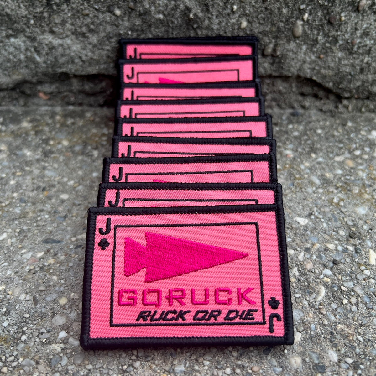 A stack of rectangular, pink morale patches for a rucking challenge featuring a black arrowhead icon, a “J” club symbol, and the text “GORUCK RUCK OR DIE.”