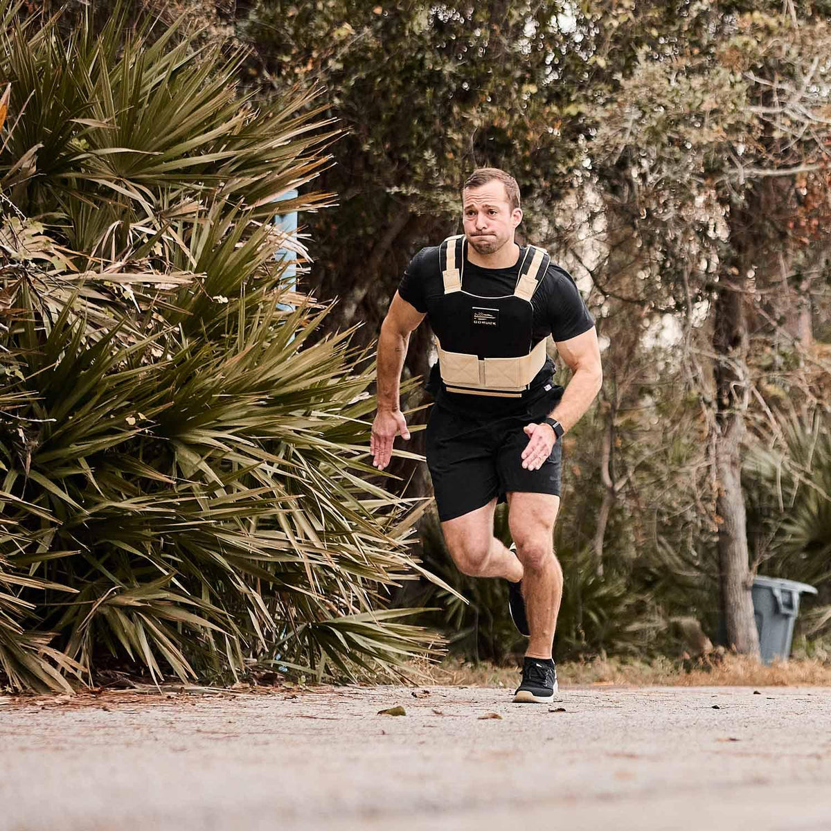 Man running outdoors in a GORUCK weighted vest and black athletic gear near greenery