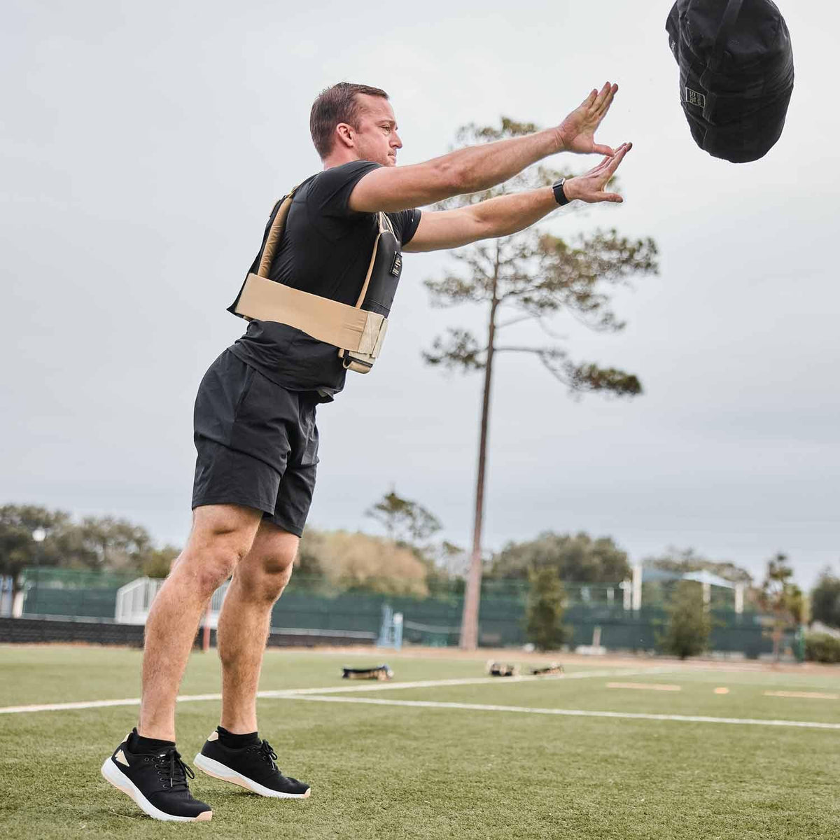 Man training outdoors in GORUCK weighted vest, tossing sandbag on athletic field