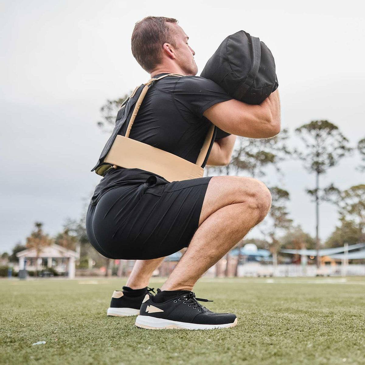 Man performing outdoor squat with GORUCK sandbag, wearing black athletic gear and trainers.
