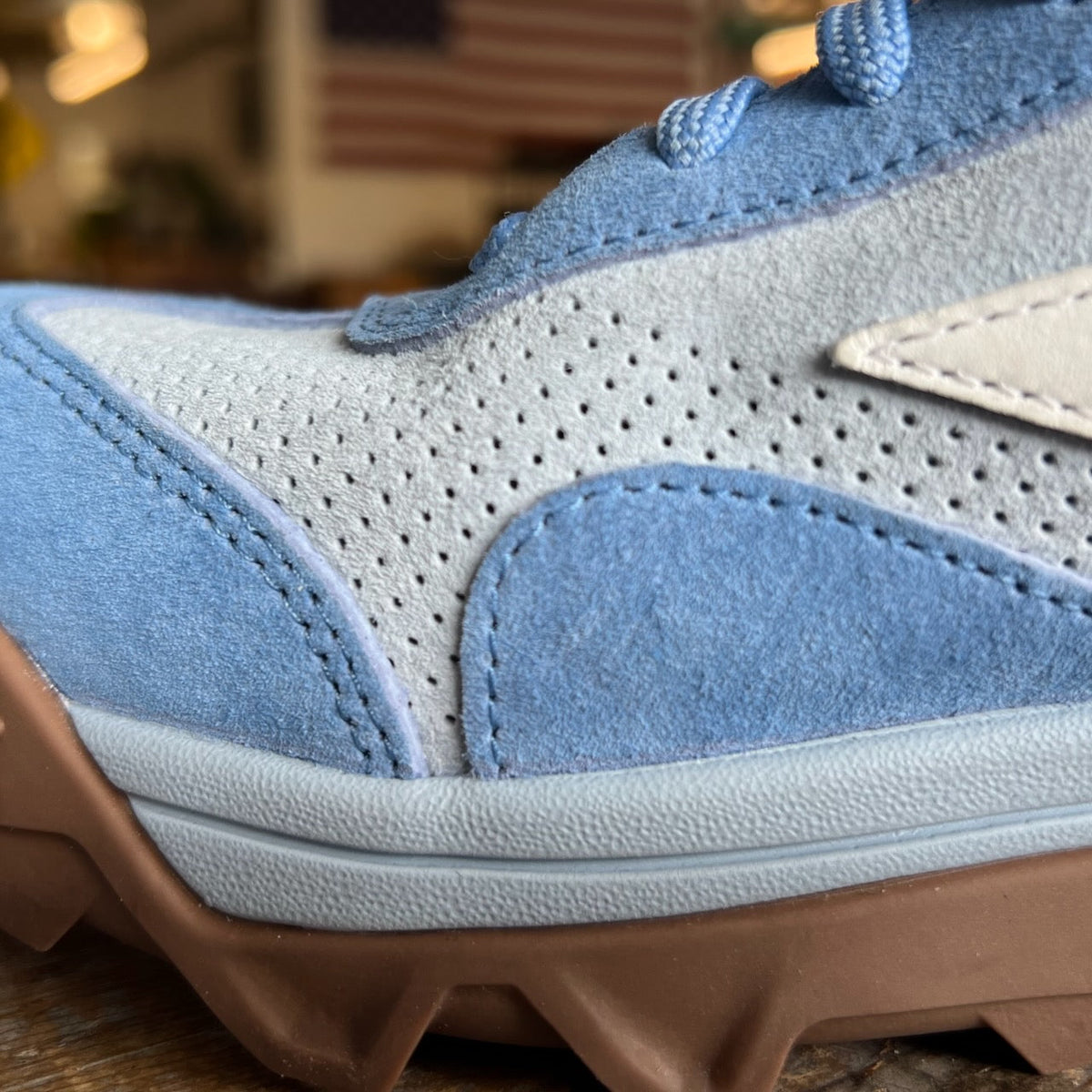 Close-up of the Mackall 45 Americana sneaker in blue and gray with a brown sole, displayed indoors against a blurred American flag backdrop.