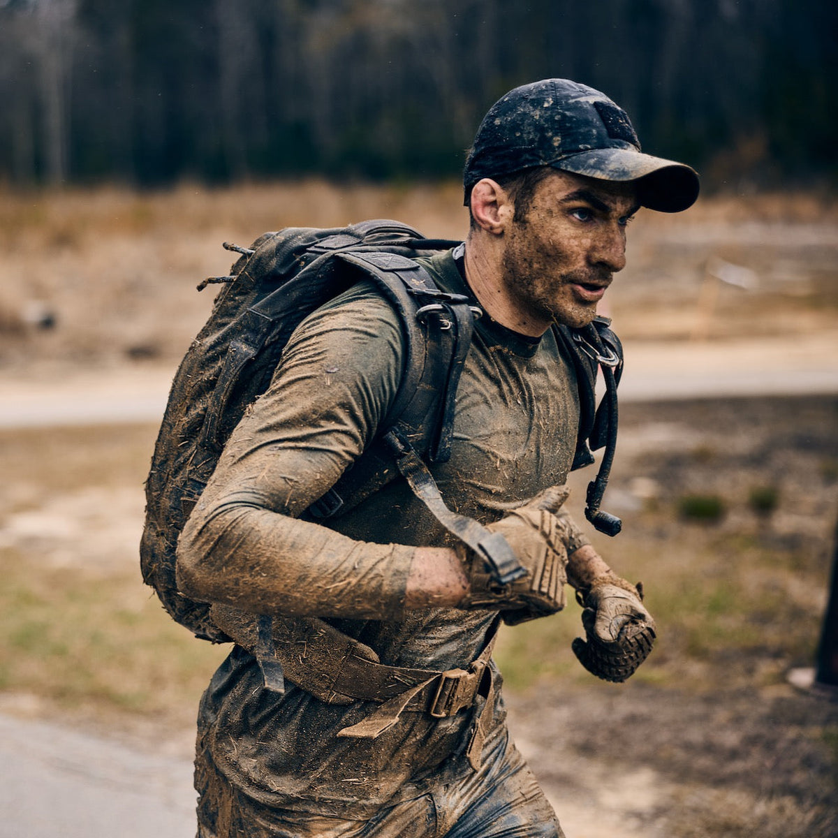 A man in a baseball cap and backpack runs through the mud, wearing abrasion resistant, tough technical pants.